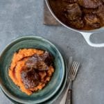 A green plate containing sweet potato puree, topped with 2 pieces of short rib. A Dutch oven is beside the bowl with more short ribs.