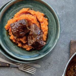 A green plate containing sweet potato puree, topped with 2 pieces of short rib. A Dutch oven is beside the bowl with more short ribs.