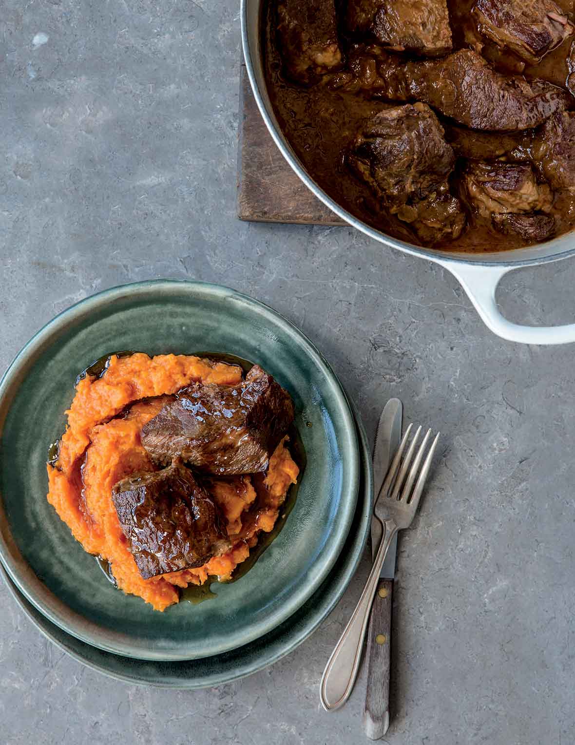A green plate containing sweet potato puree, topped with 2 pieces of short rib. A Dutch oven is beside the bowl with more short ribs.