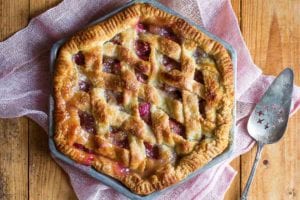 A hexagonal rhubarb pie with a lattice top crust on a pink linen cloth with a pie server resting beside the pie.