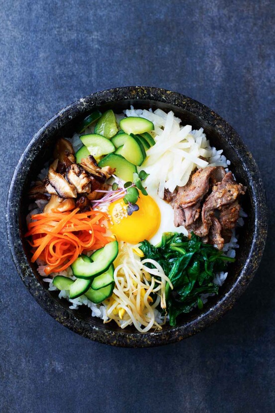 Bibimbap in an enamel bowl with sections of sprouts, noodles, spinach, carrots, meat, cucumber.