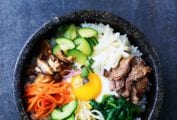 Bibimbap in an enamel bowl with sections of sprouts, noodles, spinach, carrots, meat, cucumber.