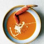A bowl of creamy crab soup drizzled with cream, and sprinkled with pepper, with a crab leg sticking out of it.
