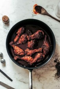 A skillet filled with chicken drumsticks braised in wine with a wooden spoon and wine cork lying beside the skillet.