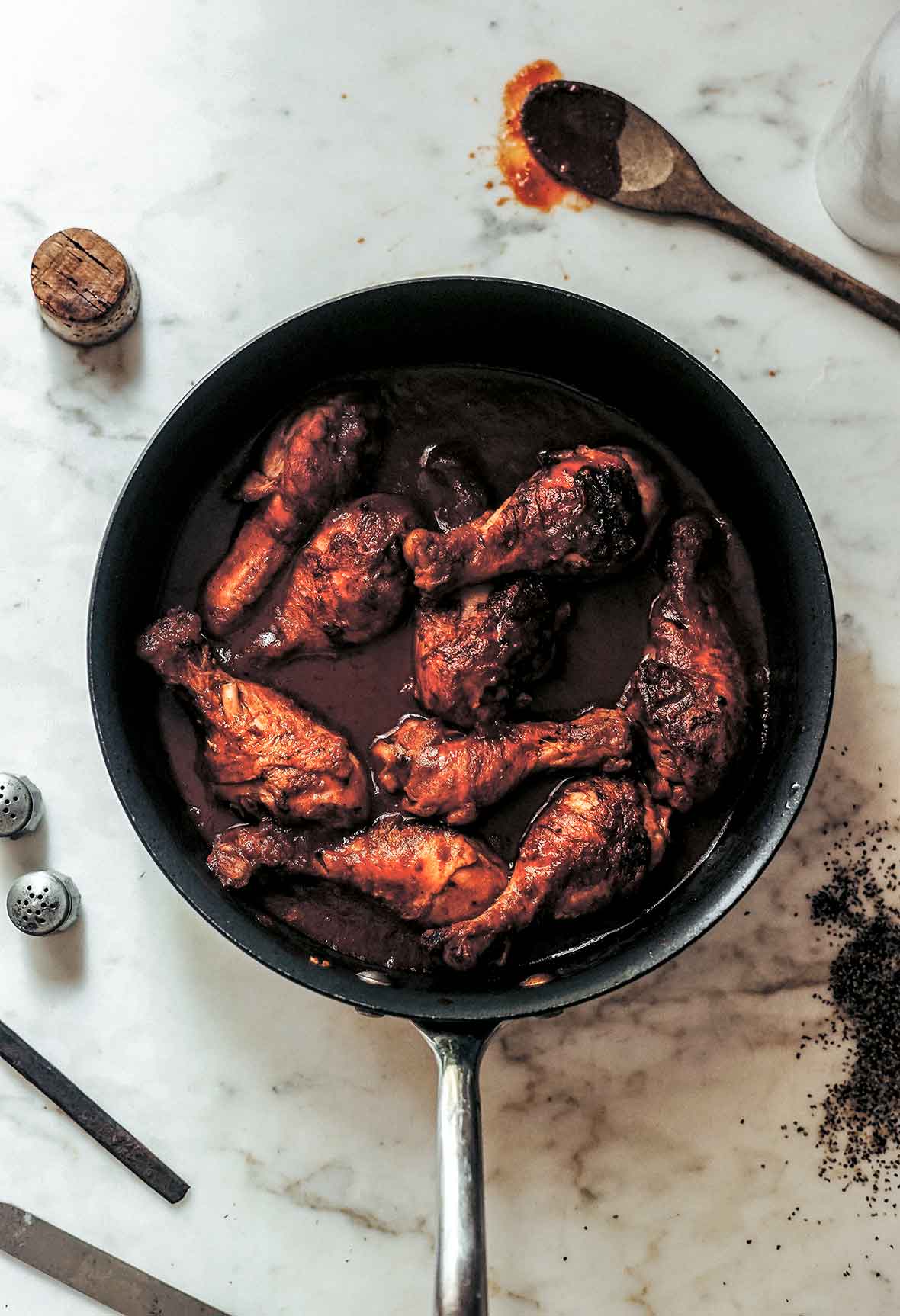 A pan filled with chicken drumsticks stewed in wine, with a wooden spoon and a wine cork next to the pan.