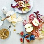 A wooden cutting board topped with halved apples, cut figs, and cut radicchio to the right of a white plate filled with the salad.