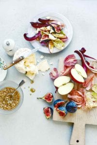 A wooden cutting board topped with halved apples, cut figs, and cut radicchio to the right of a white plate filled with the salad.
