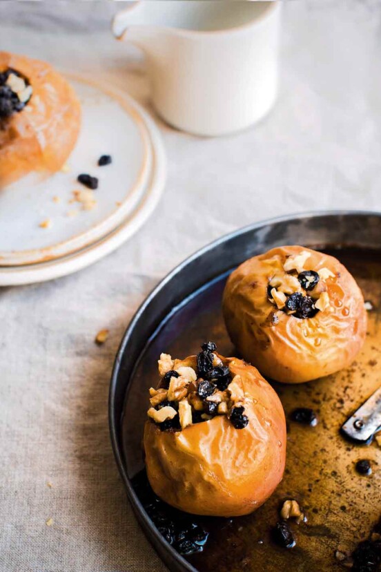 A metal baking dish with two baked apples in it, each apple topped with almonds and currants.