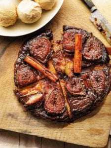 A fig tarte tatin, sliced into four pieces with cinnamon sticks on top. Nearby a bowl of ice cream.