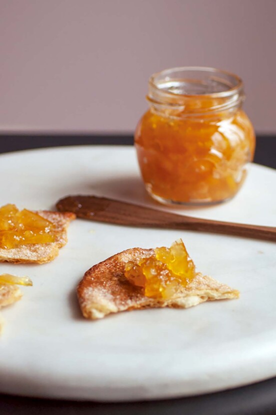 A serving platter with a piece of toast spread with marmalade. A jar of marmalade and knife rest in the background.