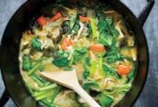 A steel wok filled with mushrooms, greens, broth, and carrots, with a wooden spoon resting inside