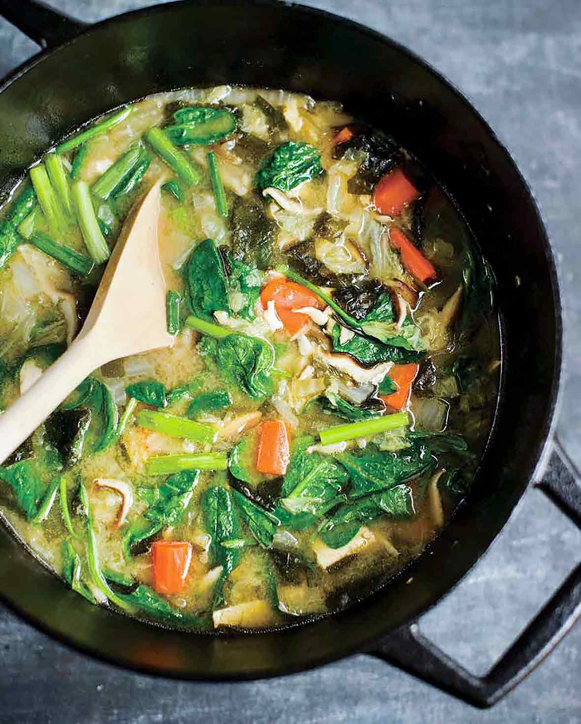 A steel wok filled with mushrooms, greens, broth, and carrots, with a wooden spoon resting inside