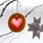A scalloped round sugar cookie with a heart-shaped windowpane cutout hanging from a tree branch.