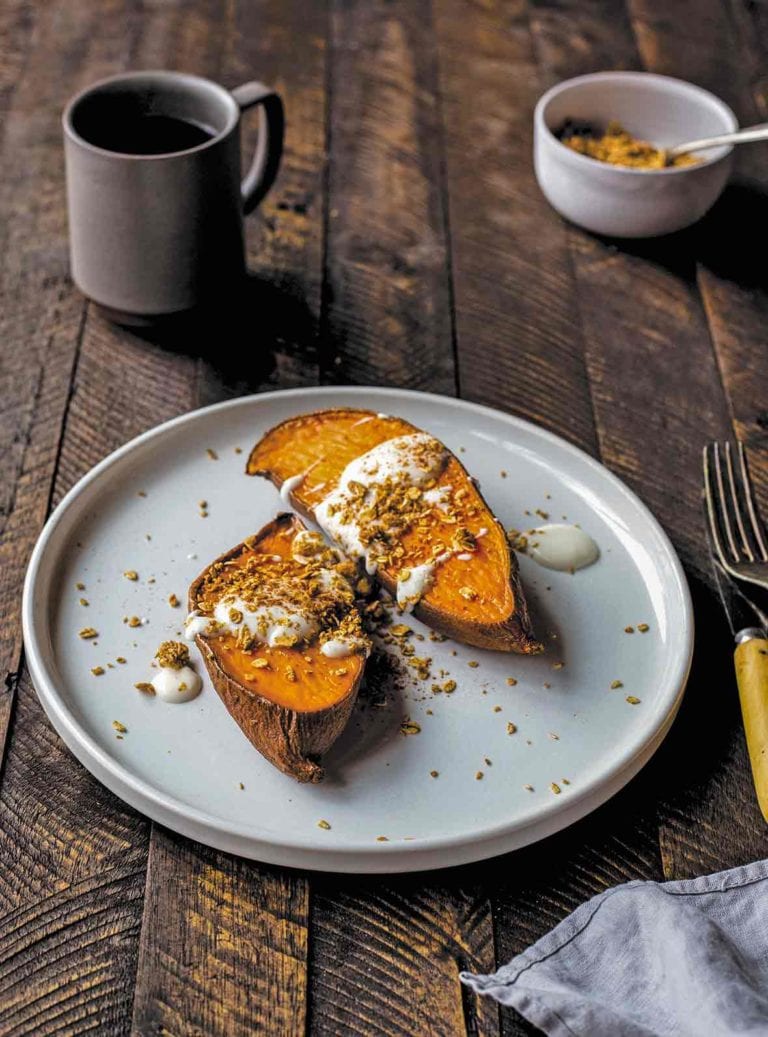 Two halves of a sweet potato drizzled with maple-yogurt sauce and topped with granola on a plate on a wooden table