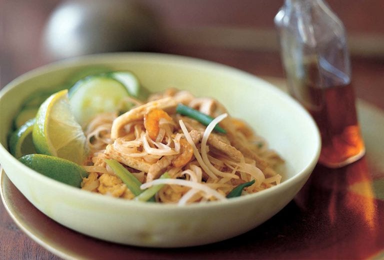 Pad Thai noodles, crunchy noodles. cucumber, bean sprouts, and lime wedges in a bowl, a bottle of chile-vinegar