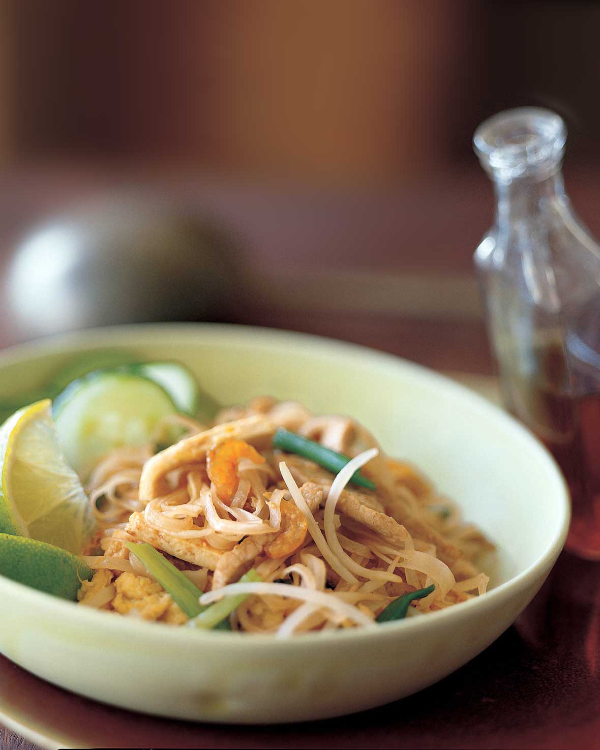 Pad Thai noodles, crunchy noodles. cucumber, bean sprouts, and lime wedges in a bowl, a bottle of chile-vinegar