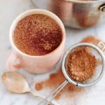 A pot and a mug filled with hot cocoa and a sifter of cocoa powder on the side.