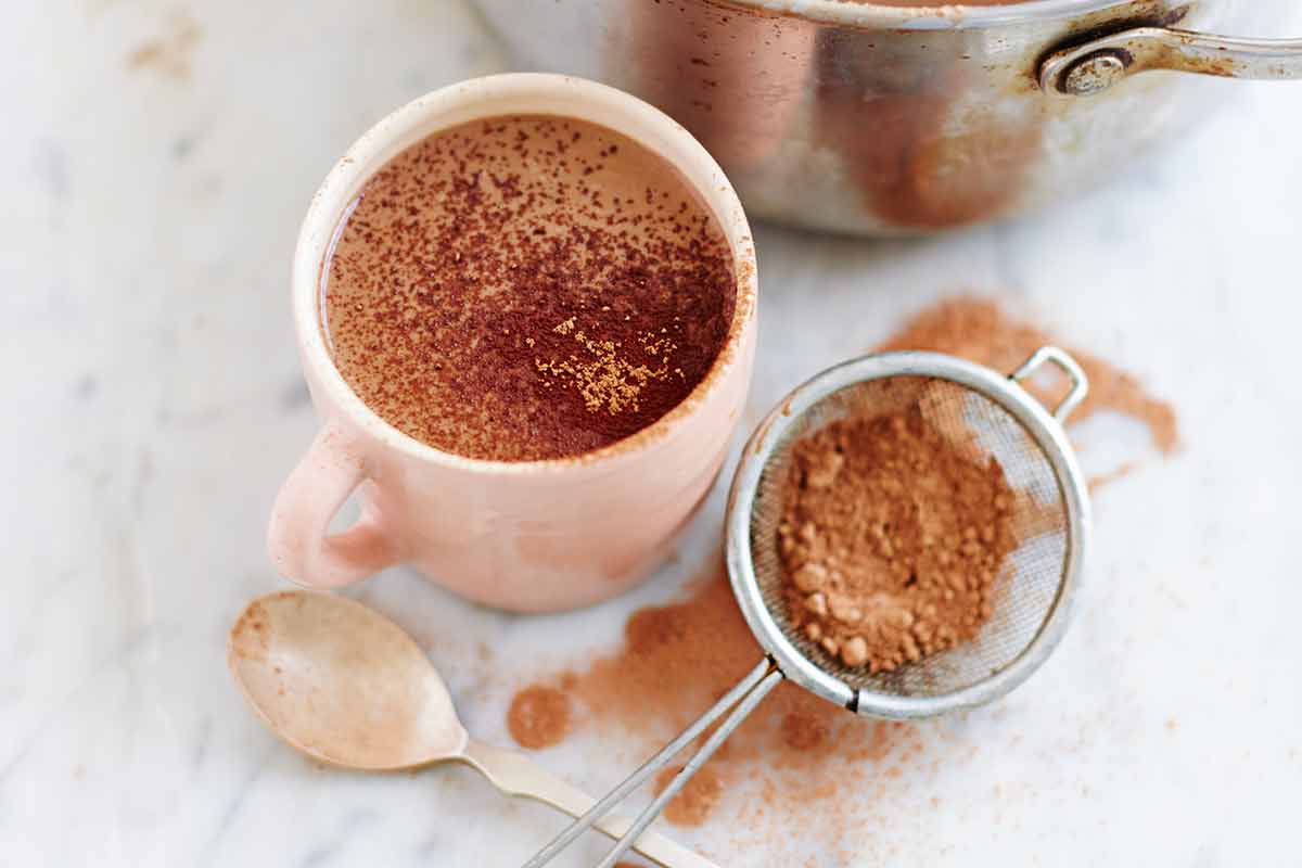 A pot and a mug filled with hot cocoa and a sifter of cocoa powder on the side.