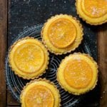Five small tarts filled with orange curd and topped with candied orange slices on a cooling rack