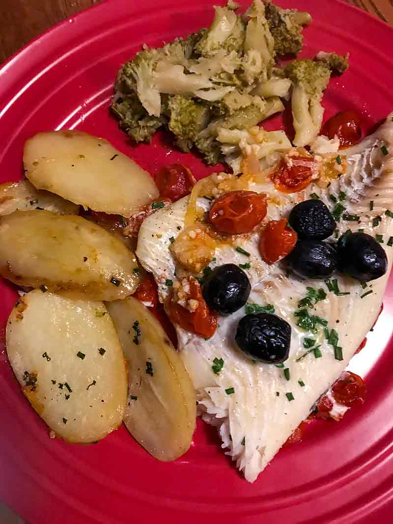 Red plate with baked fish with tomatoes and olives, roasted potatoes, broccoli