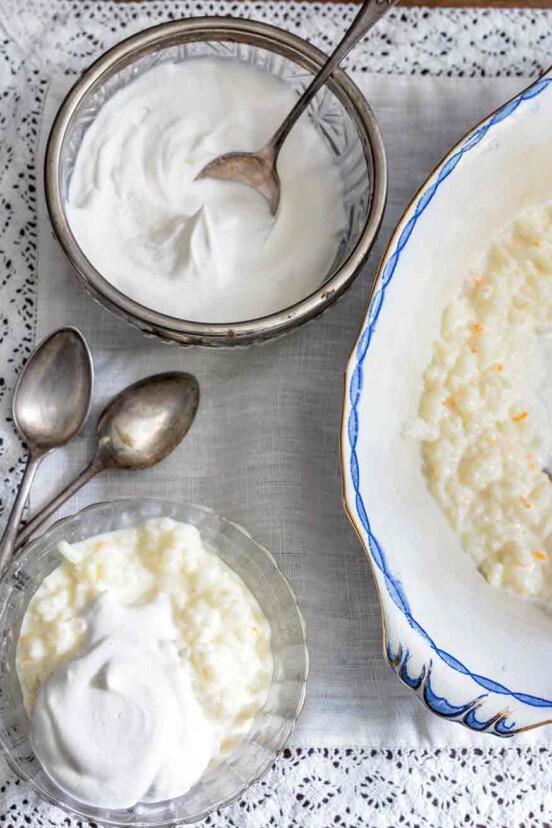 Serving bowl and small bowl of French rice pudding topped with whipped cream, along with spoons on a lace tablecloth.