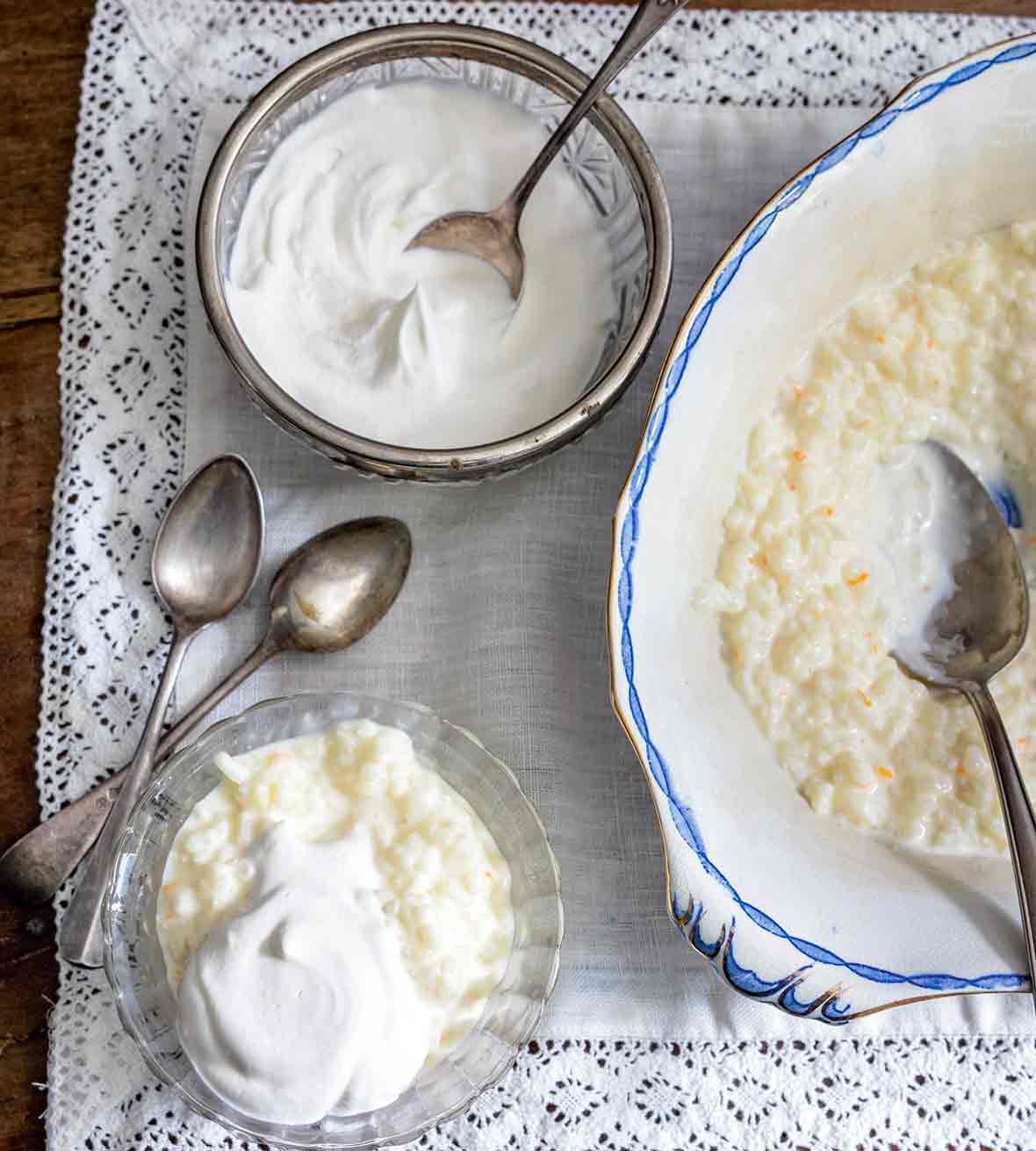Serving bowl and small bowl of French rice pudding topped with whipped cream, along with spoons on a lace tablecloth.