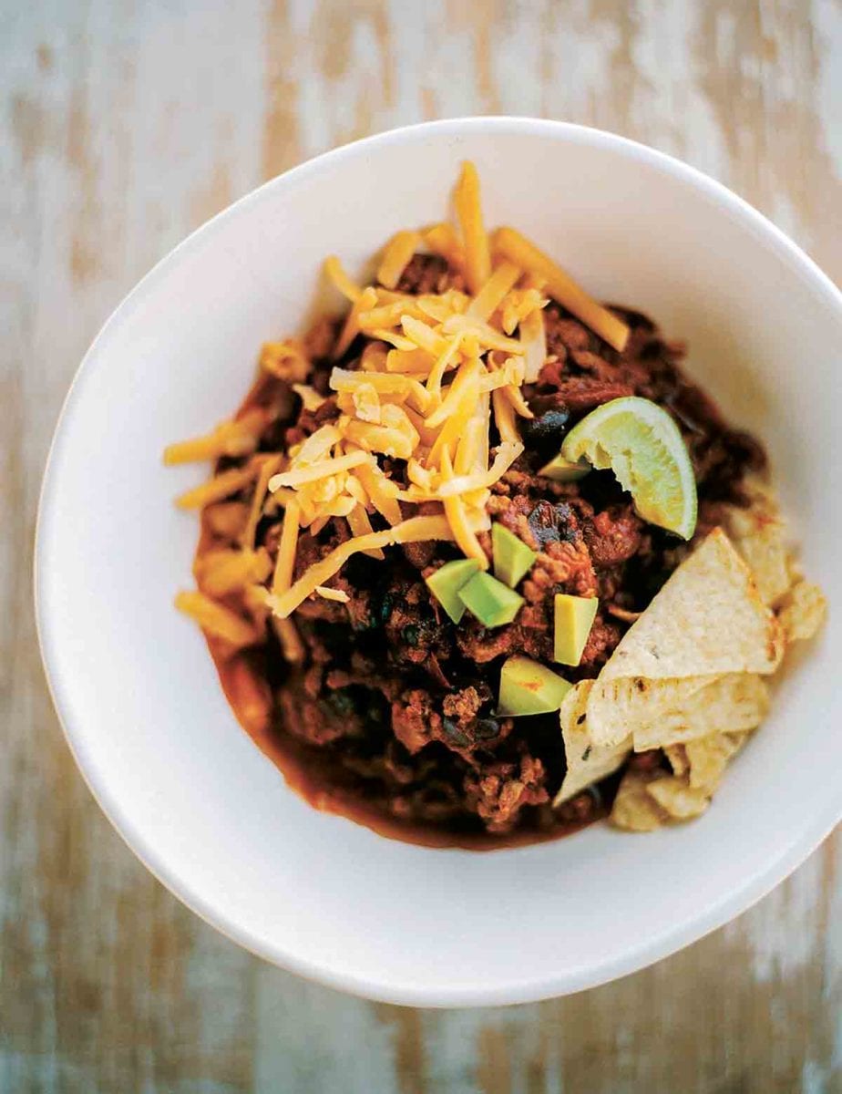 White bowl of beef chili with grated cheese, tortilla chips and lime wedges on wood