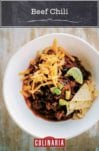 White bowl of beef chili with grated cheese, tortilla chips and lime wedges on wood