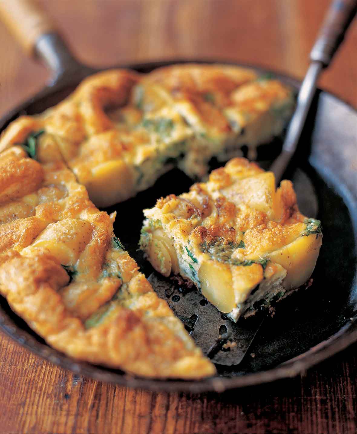 Cast iron skillet of broccolini and potato frittata, with a wedge on a metal spatula