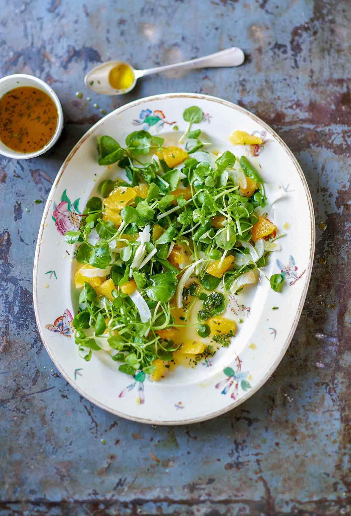 Plate of fennel, orange, and watercress salad on a metal sheet with dressing and a spoon on the side