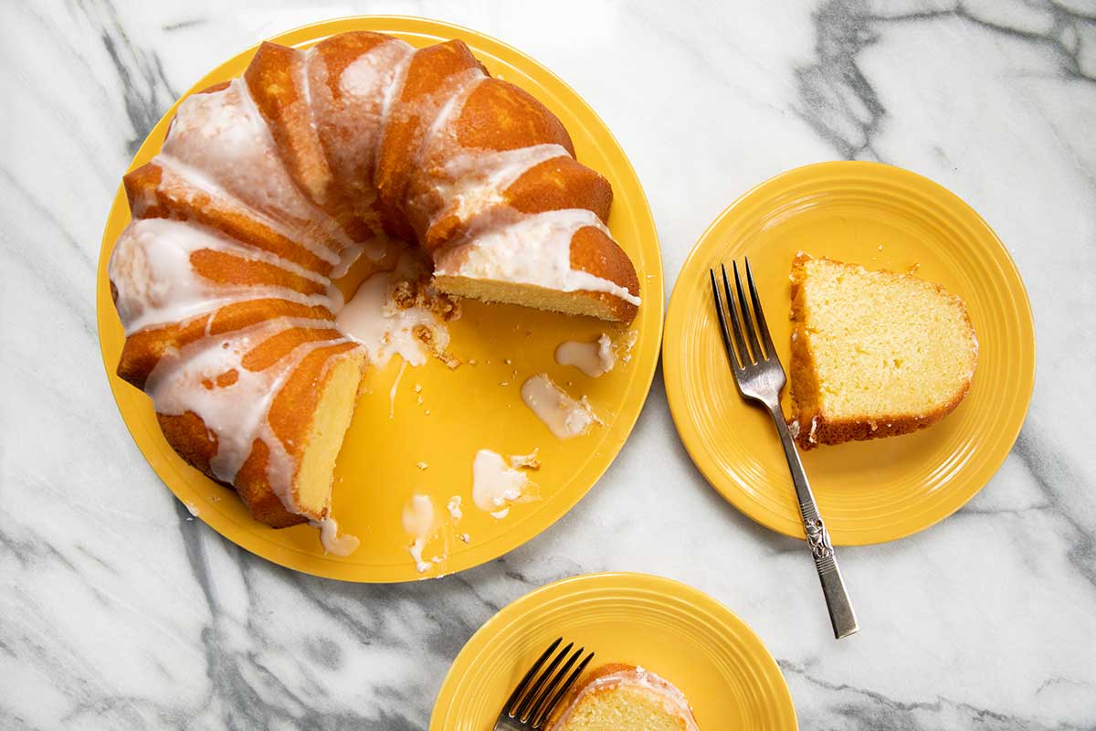 A partially cut lemon pound cake on a yellow platter with slices and forks on two yellow plates.
