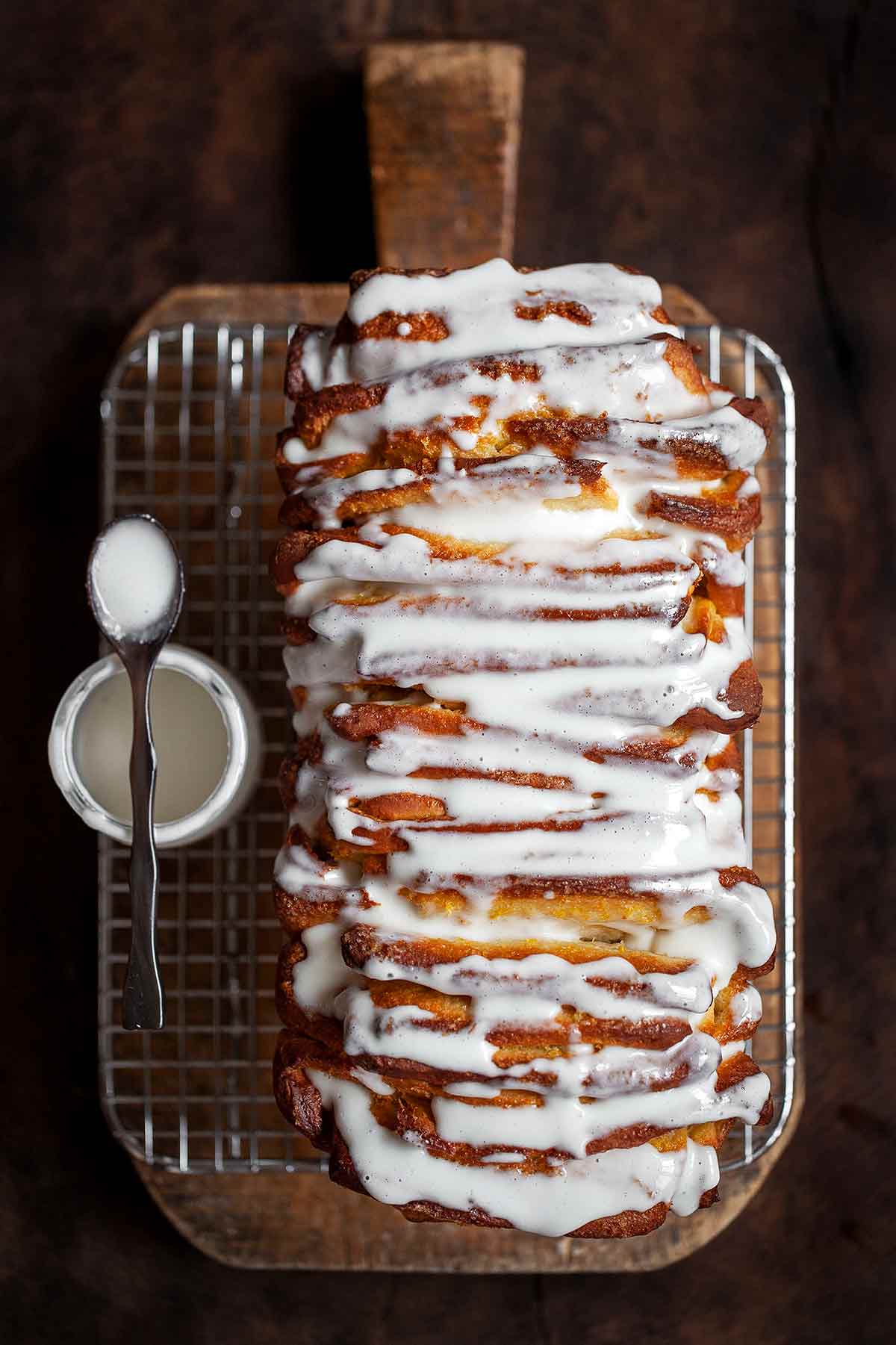 A glazed lemon pull-apart coffee cake on a wire rack with a spoon and jar of glaze beside it, all resting on a wooden cutting board.