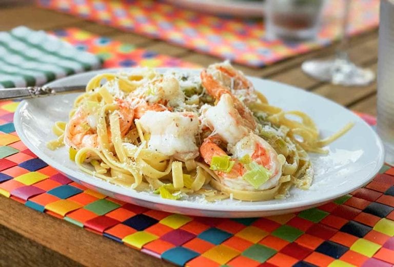 A white plate filled with shrimp and leek pasta on a multi-colored woven placemat.