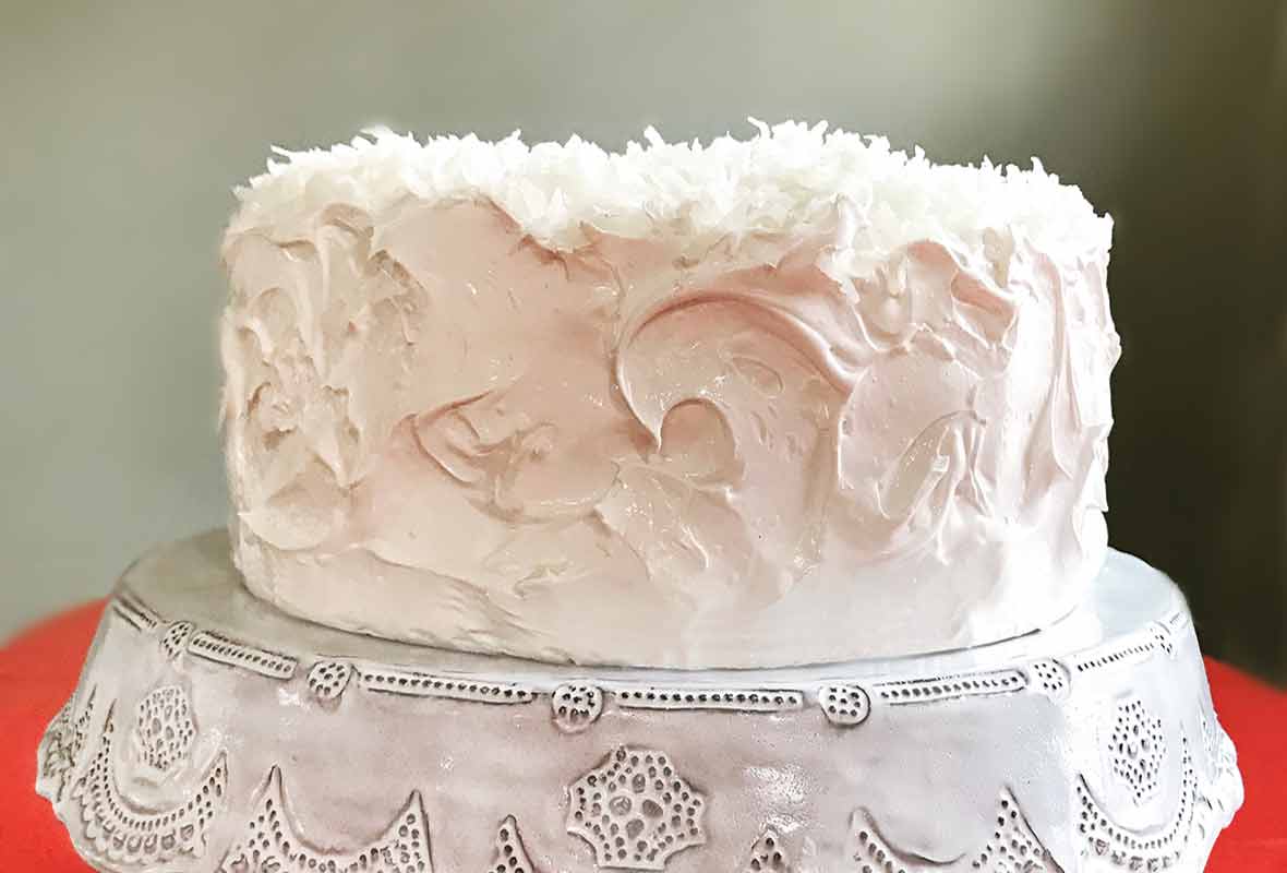 Coconut layer cake with white meringue frosting topped with shredded coconut on a white cake stand