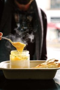 Person with a spoon digging into a glass jar of coddle eggs with potato puree underneath, slice of bread nearby