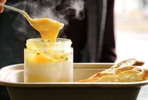 Person with a spoon digging into a glass jar of coddle eggs with potato puree underneath, slice of bread nearby