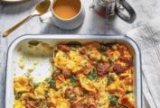 Baking dish of sausage egg casserole of bread, eggs, sausage, tomato, kale, and Cheddar cheese, a plate and tea nearby
