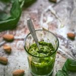 Glass of spinach almond pesto with a spoon inside on a metal sheet strewn with almonds