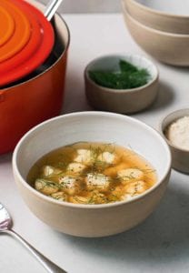 A bowl of chicken soup with dumplings, topped with fennel fronds, along with red pot and bowls