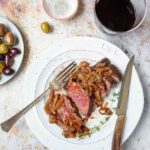 A white plate with a sliced grilled bavette steak with caramelized shallots on top, a glass of wine and olives nearby