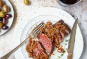 A white plate with a sliced grilled bavette steak with caramelized shallots on top, a glass of wine and olives nearby