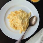 A pile of perfect scrambled eggs on a white plate with a spoon resting beside the eggs.