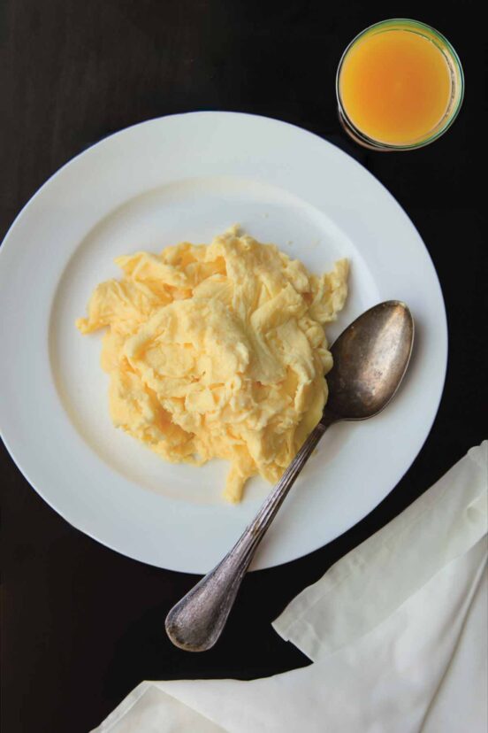 A pile of perfect scrambled eggs on a white plate with a spoon resting beside the eggs.