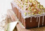 A pistachio lover's pound cake on a wooden cutting board with a mesh bag of pistachios beside it.