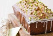 A pistachio lover's pound cake on a wooden cutting board with a mesh bag of pistachios beside it.