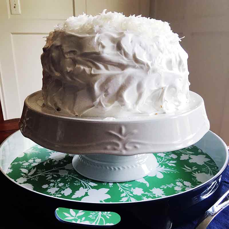 A Magnolia Bakery coconut cake--with meringue frosting and topped with coconut on a white cake stand, on a green tray