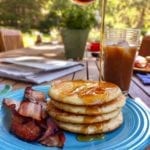 A blue plate topped with three light and fluffy pancakes, maple syrup, and cooked bacon.