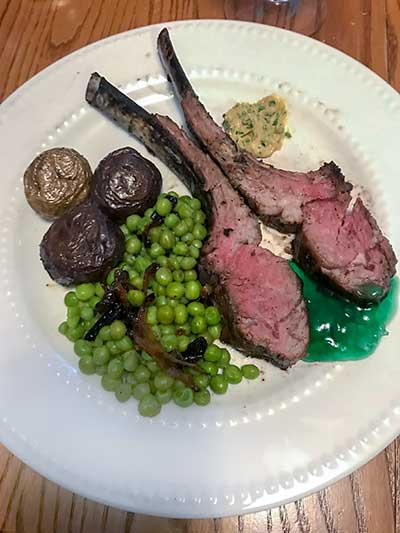 White plate with roasted rack of lamb with parsley, dijon, and chives, peas, potatoes, and mint jelly