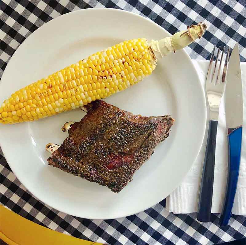A rack of pressure cooker ribs on a white plate with an ear of corn, all on a blue-and-white checked tablecloth