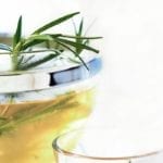 A pot of rosemary infused tea with rosemary sprigs sticking out the top and a glass beside it.